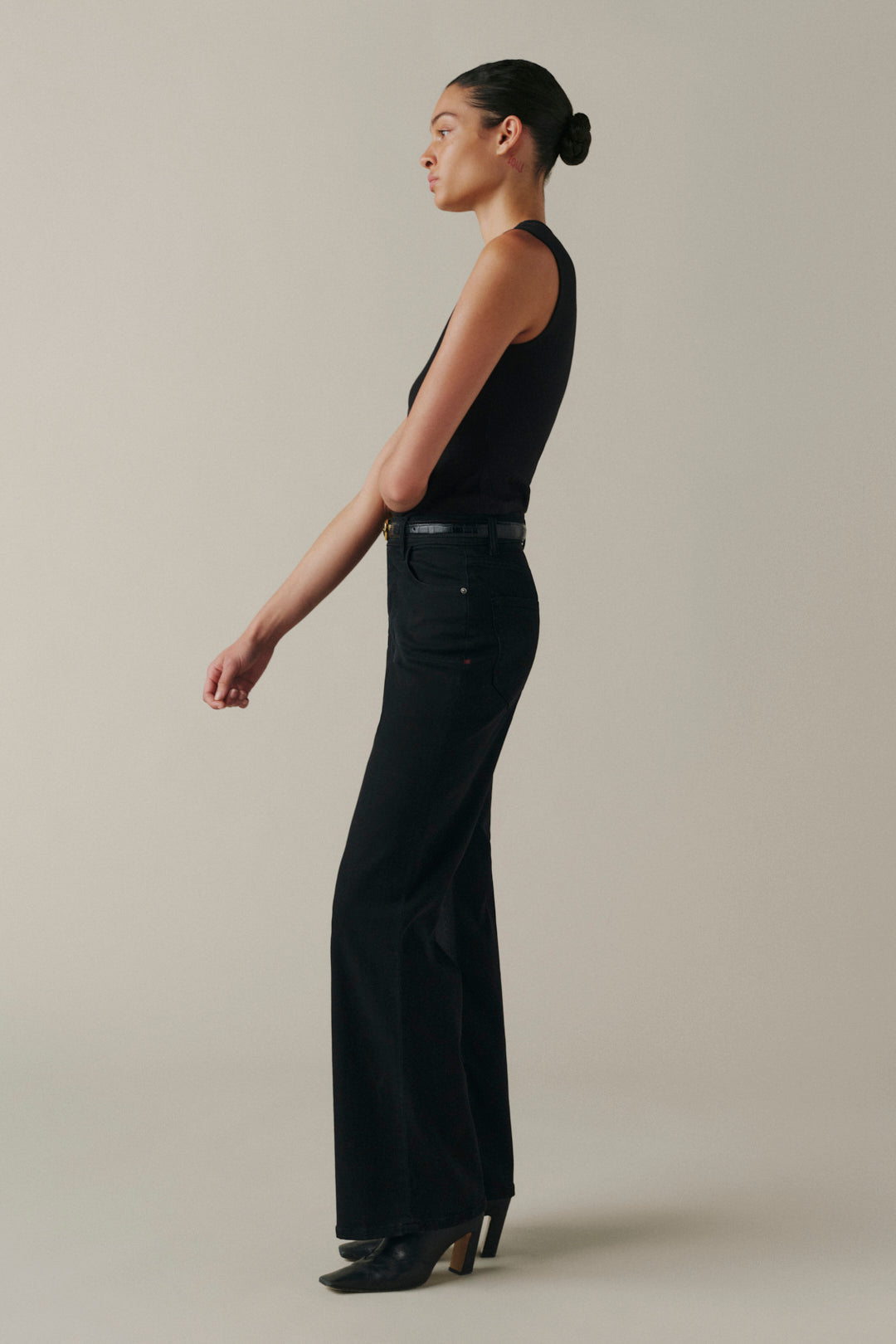A woman in a black sleeveless top and black wide-leg trousers stands in profile against a plain warm beige studio background.