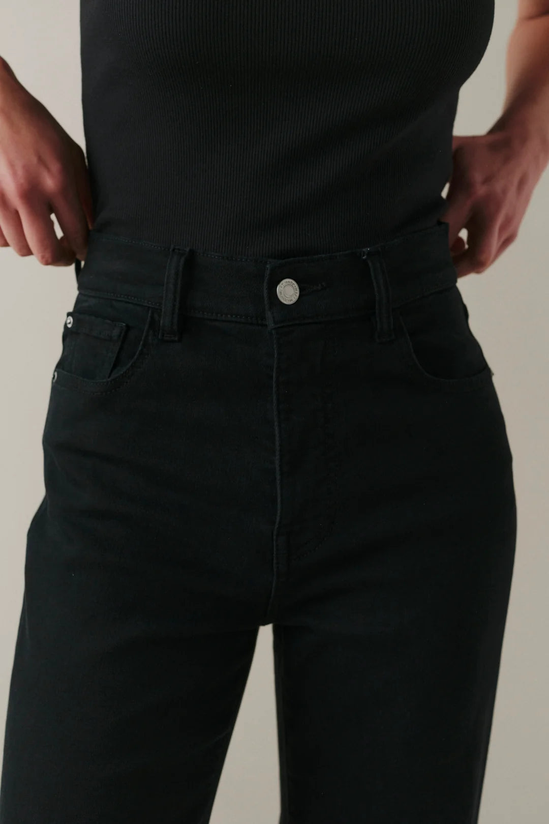Close-up of a person wearing black high-rise jeans with a silver button closure, paired with a black ribbed top, against a light beige background.