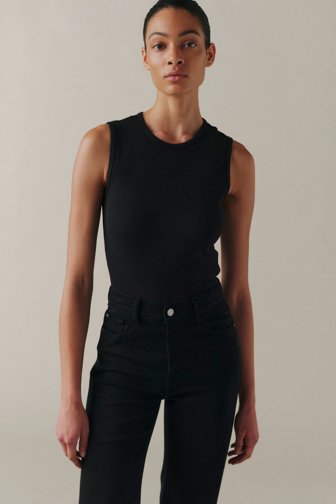 A model wearing a black sleeveless crewneck bodysuit paired with black jeans, standing against a neutral beige studio background.