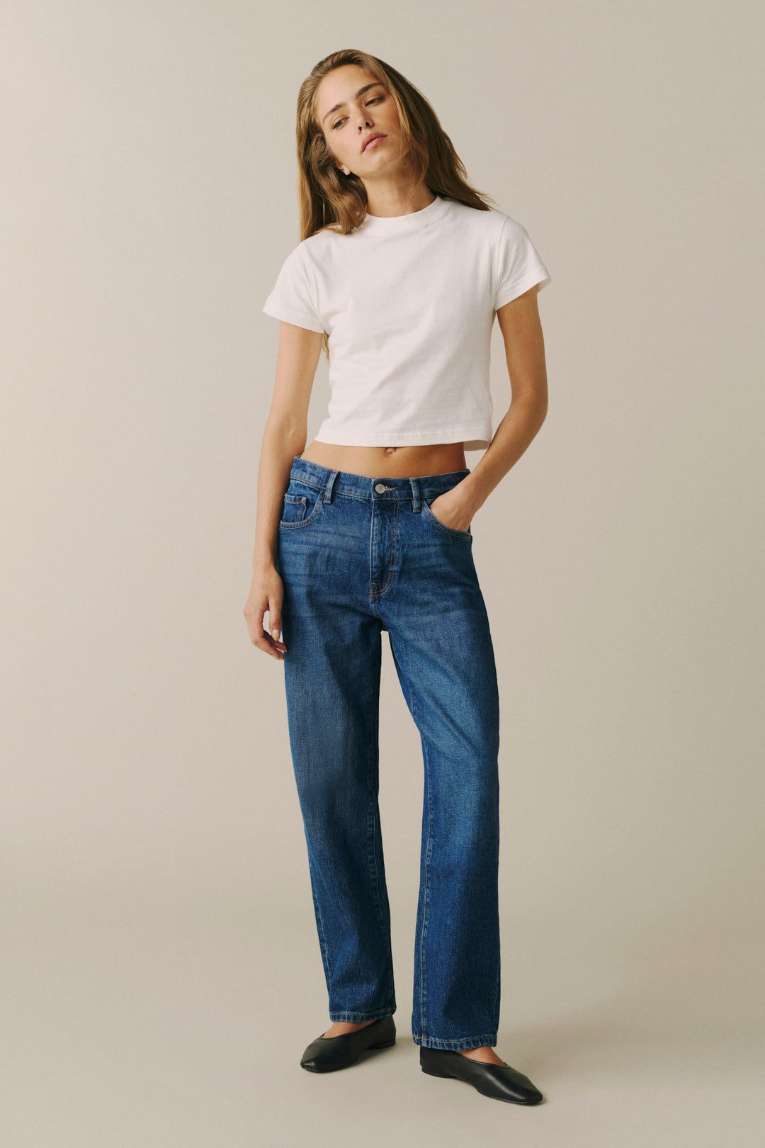 A woman modeling a white cropped short-sleeve t-shirt and straight-leg blue jeans against a solid beige studio background.