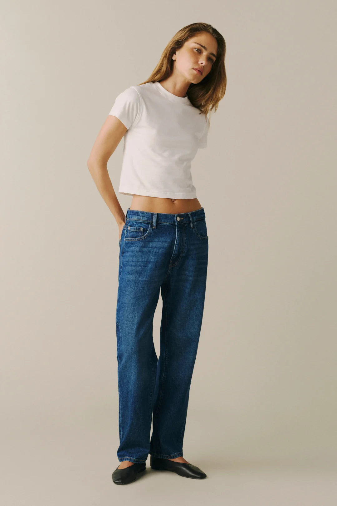 A woman modeling a pair of straight-leg blue jeans with a white cropped t-shirt and black flats.