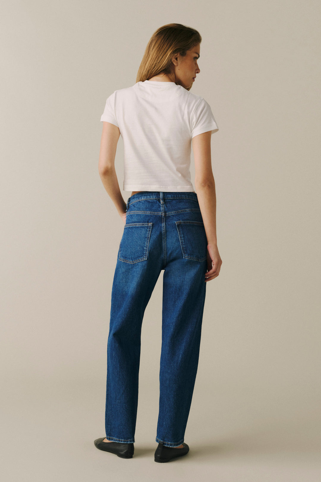 A woman shown from the back wearing blue straight-leg jeans, a white short-sleeve crop top, and black flats.