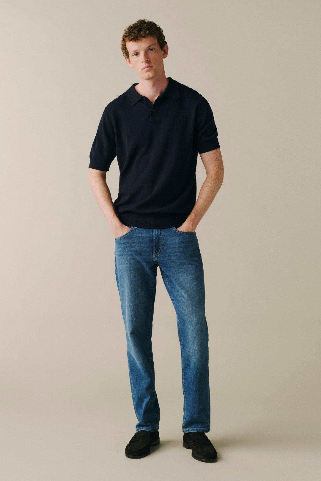 A man wearing a dark navy short-sleeve polo shirt, blue jeans, and black slip-on shoes, standing against a solid beige studio background.