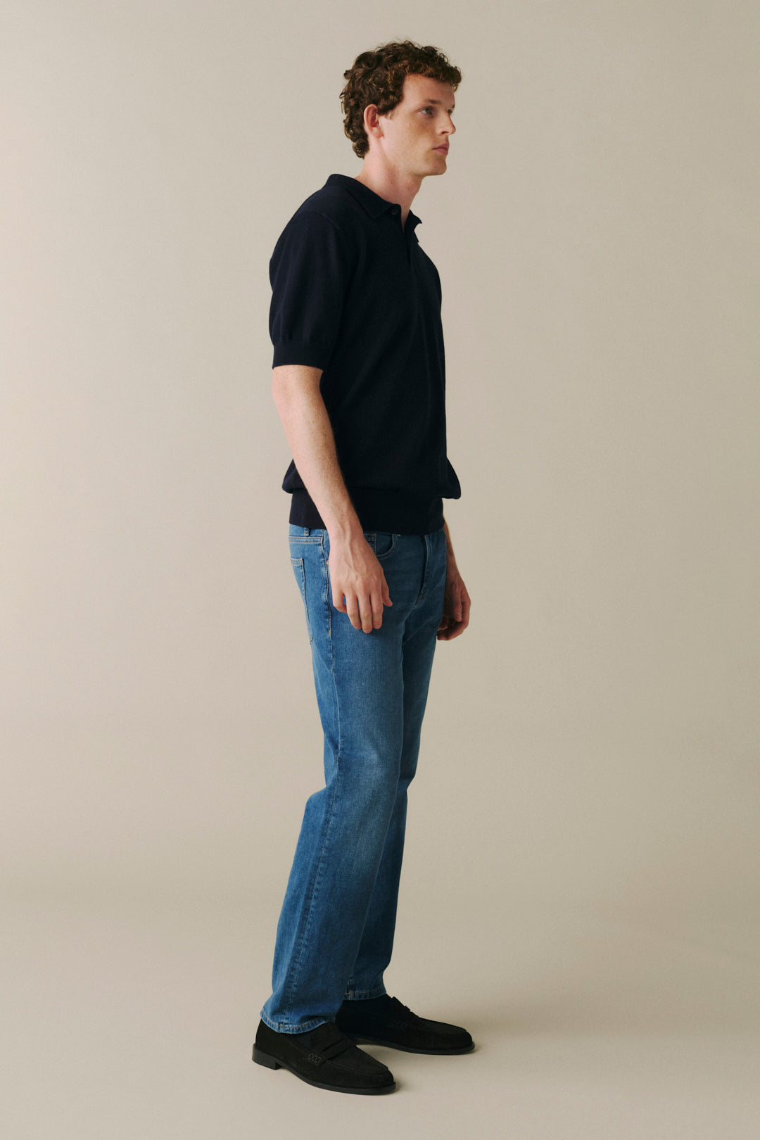 A man standing in profile wearing a dark navy short-sleeve knit polo shirt, blue jeans, and black shoes against a plain warm beige studio background.