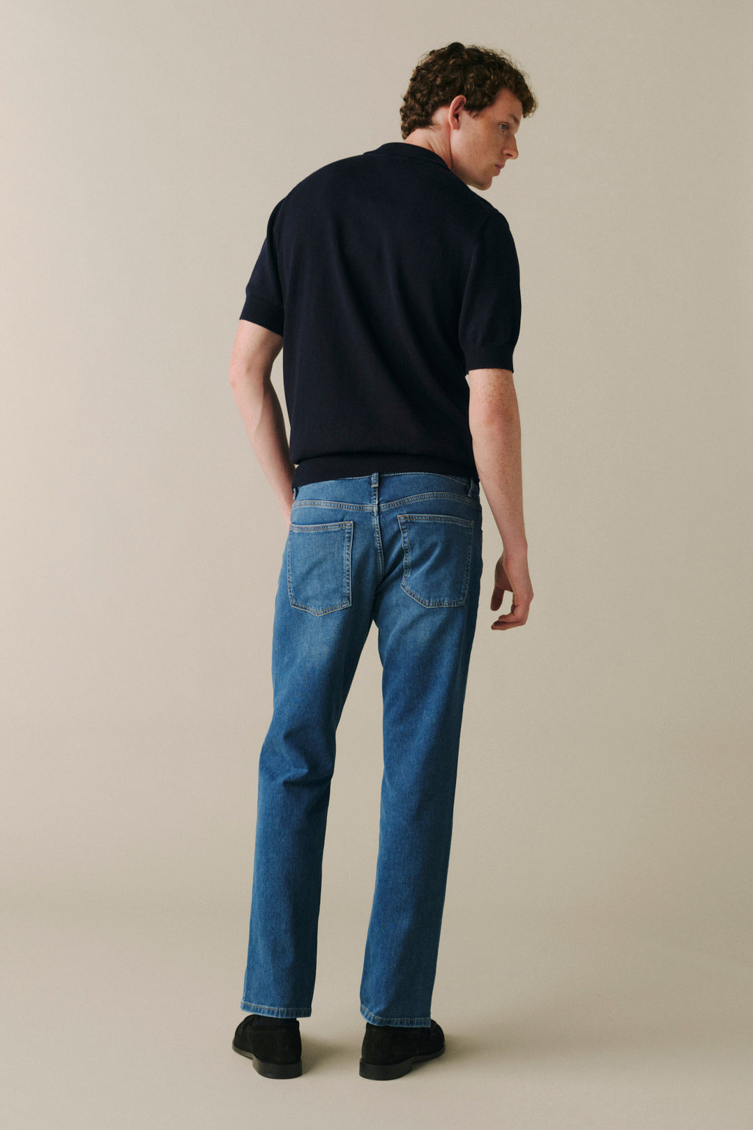 A man shown from the back wearing a dark navy short-sleeve t-shirt and blue jeans, standing against a plain beige studio background.