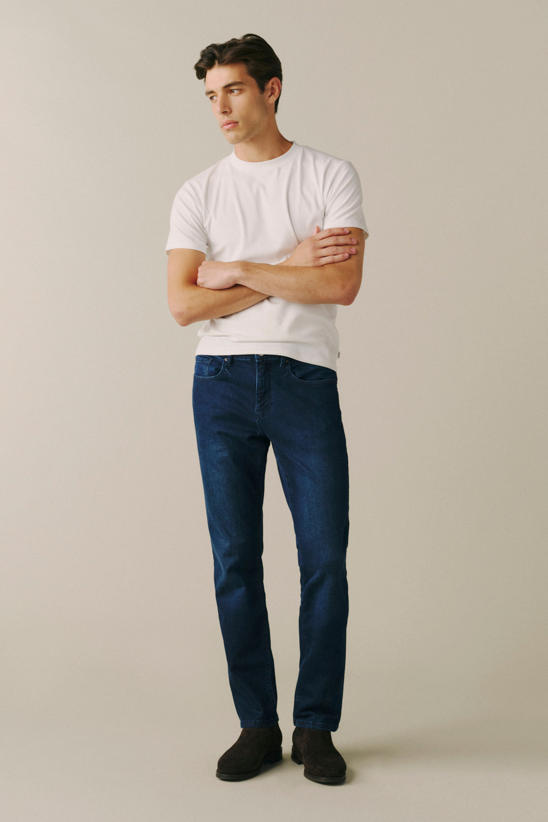A man modeling a white crewneck t-shirt and dark blue slim-fit jeans, standing with arms crossed against a solid light beige background.