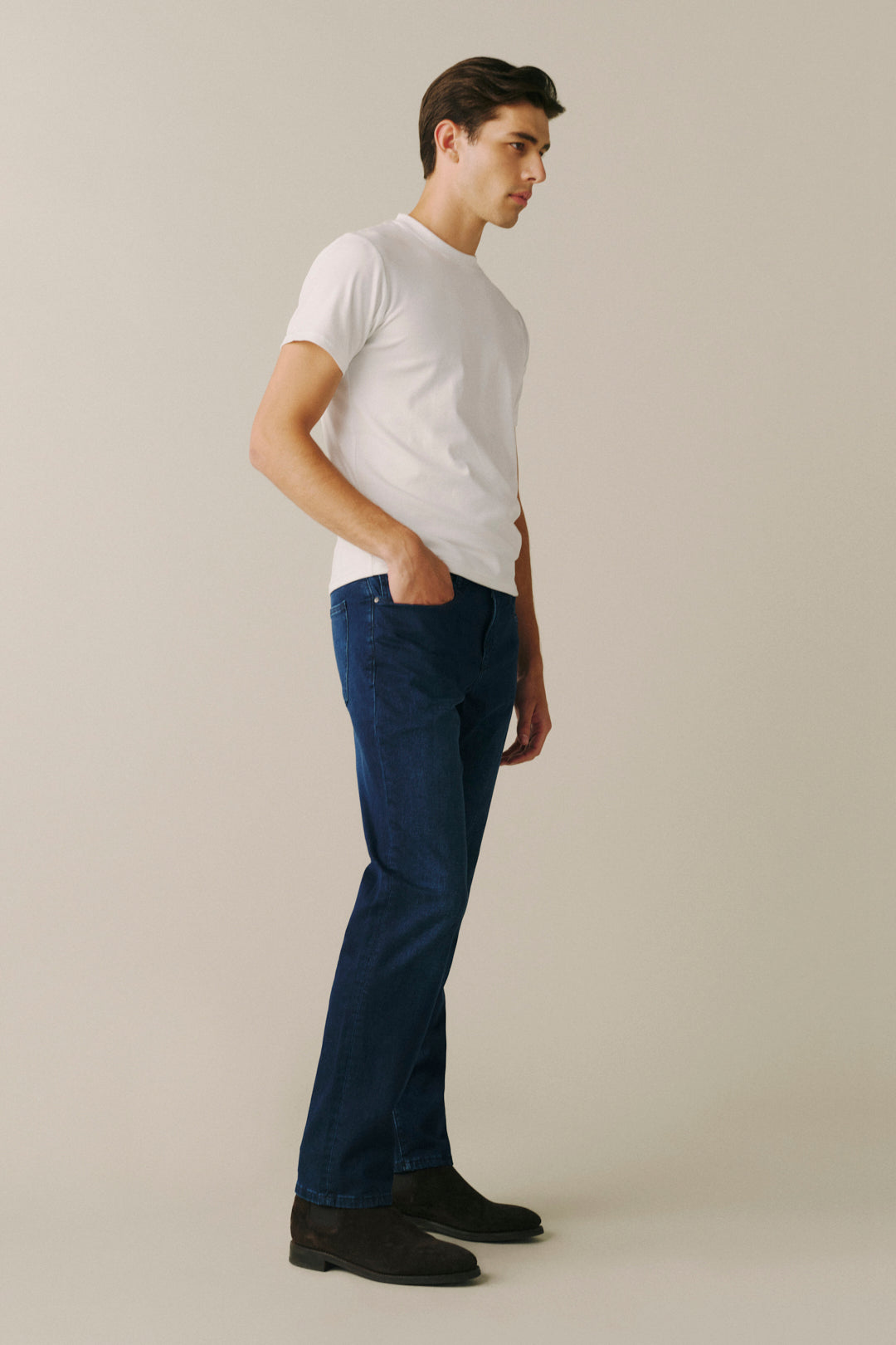 A man in a white t-shirt and dark blue jeans standing in profile against a light beige studio background.