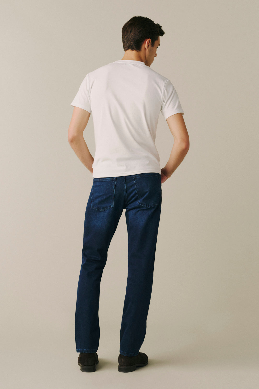 A man viewed from behind wearing a white crew-neck t-shirt, dark blue jeans, and black shoes, standing against a solid light beige studio background.