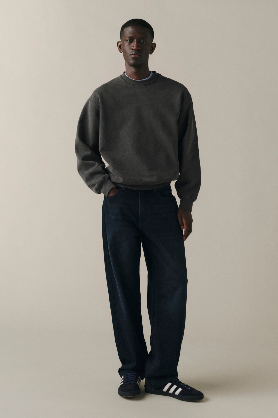 A man modeling a dark gray crewneck sweatshirt and dark navy wide-leg trousers, standing against a solid light beige studio background.