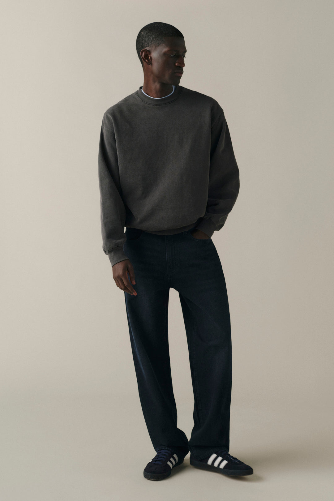 A man wearing a dark charcoal crewneck sweatshirt, dark navy wide-leg jeans, and black slide shoes with white stripes, standing against a solid light beige studio background.
