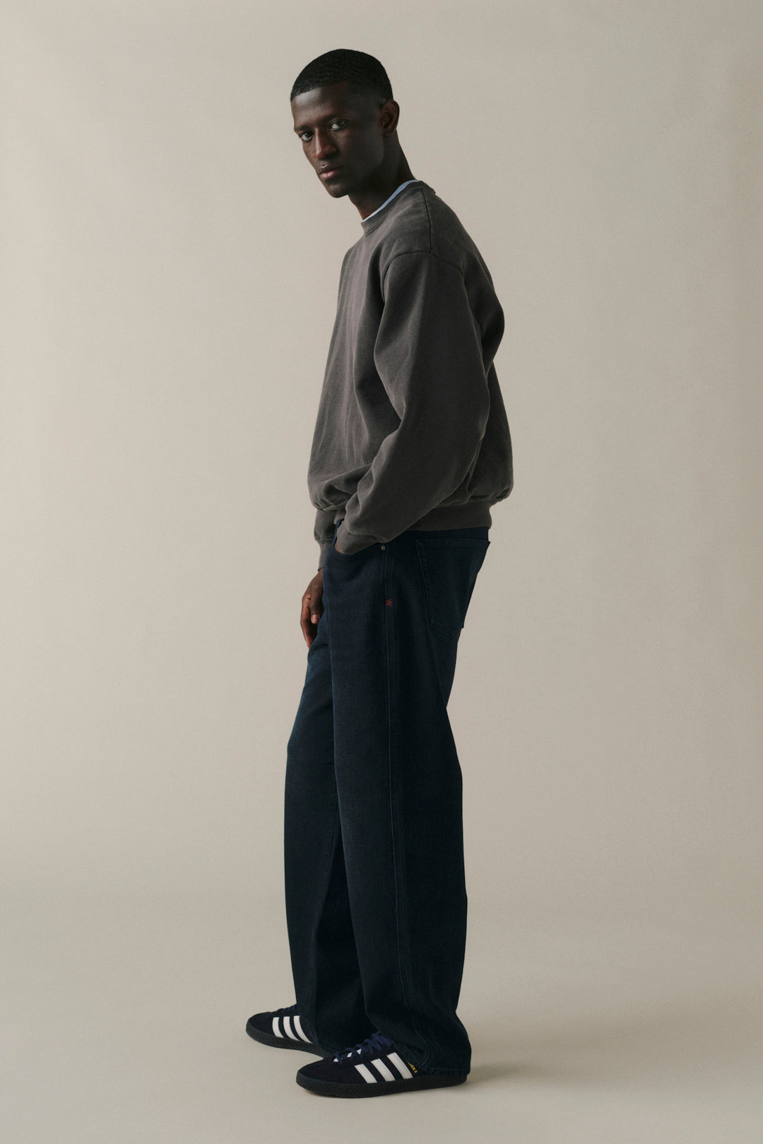 A man stands in profile wearing a dark gray crewneck sweatshirt, dark navy wide-leg trousers, and black Adidas slip-on shoes with white stripes, against a plain beige studio background.