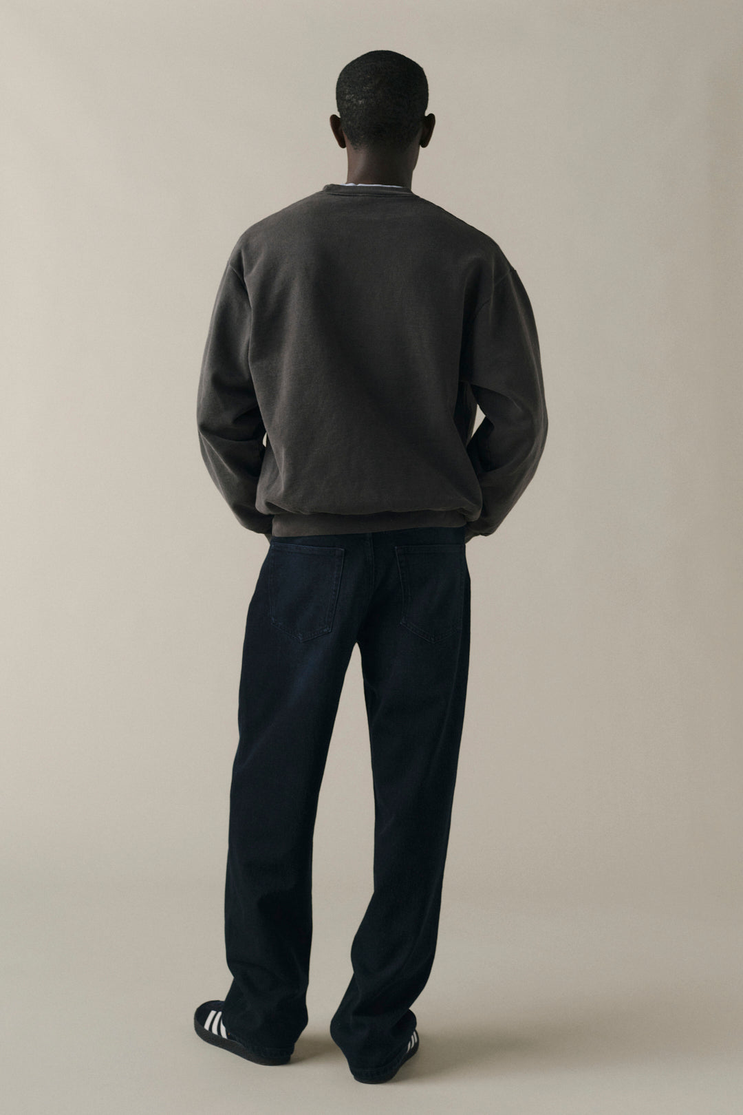 A man viewed from behind wearing a dark charcoal gray crewneck sweatshirt, dark navy pants, and black sneakers with white stripes, standing against a solid light beige background.