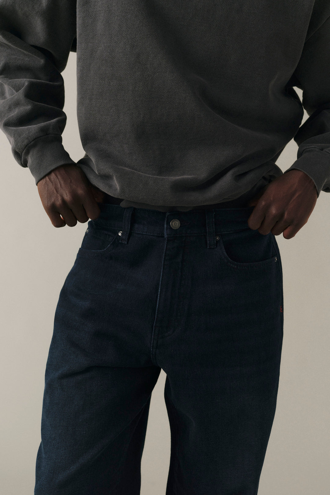 A close-up of a person wearing dark indigo blue jeans and a charcoal gray sweatshirt, standing with hands in pockets against a plain light beige background.