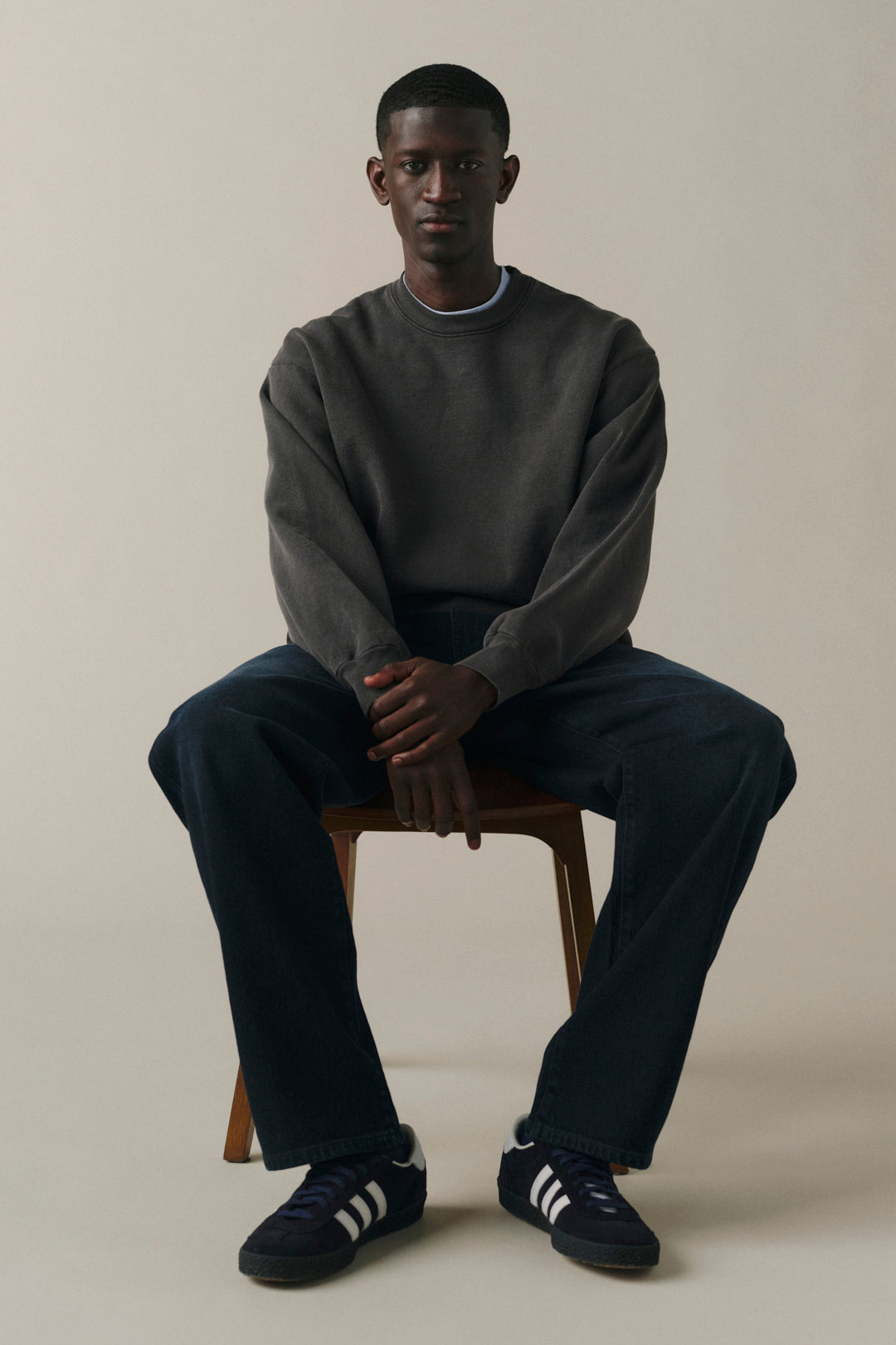 A man sits on a wooden stool wearing a dark gray crewneck sweatshirt, dark blue jeans, and navy blue sneakers with white stripes, against a plain beige studio background.