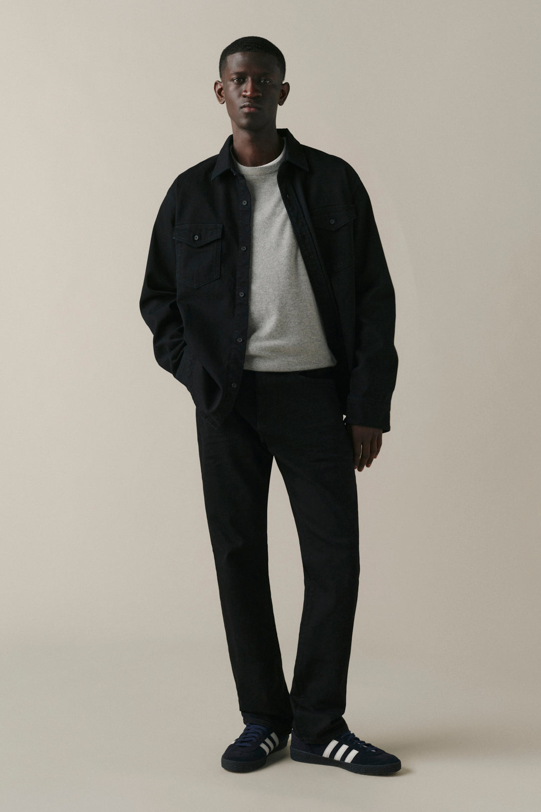 A man modeling a dark navy blue overshirt, black trousers, a light gray knit sweater, and navy blue Adidas sneakers with white stripes, standing against a solid beige studio background.