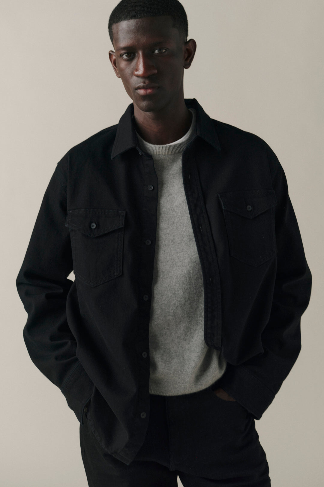 A man wearing a black overshirt jacket over a light gray crewneck sweater and black trousers, standing against a plain beige studio background.