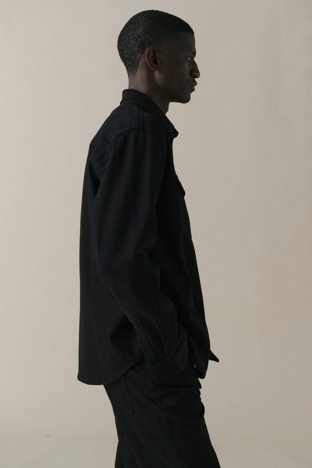 A man in profile wearing a black long-sleeve button-up shirt and matching black trousers, standing against a plain light beige studio background.