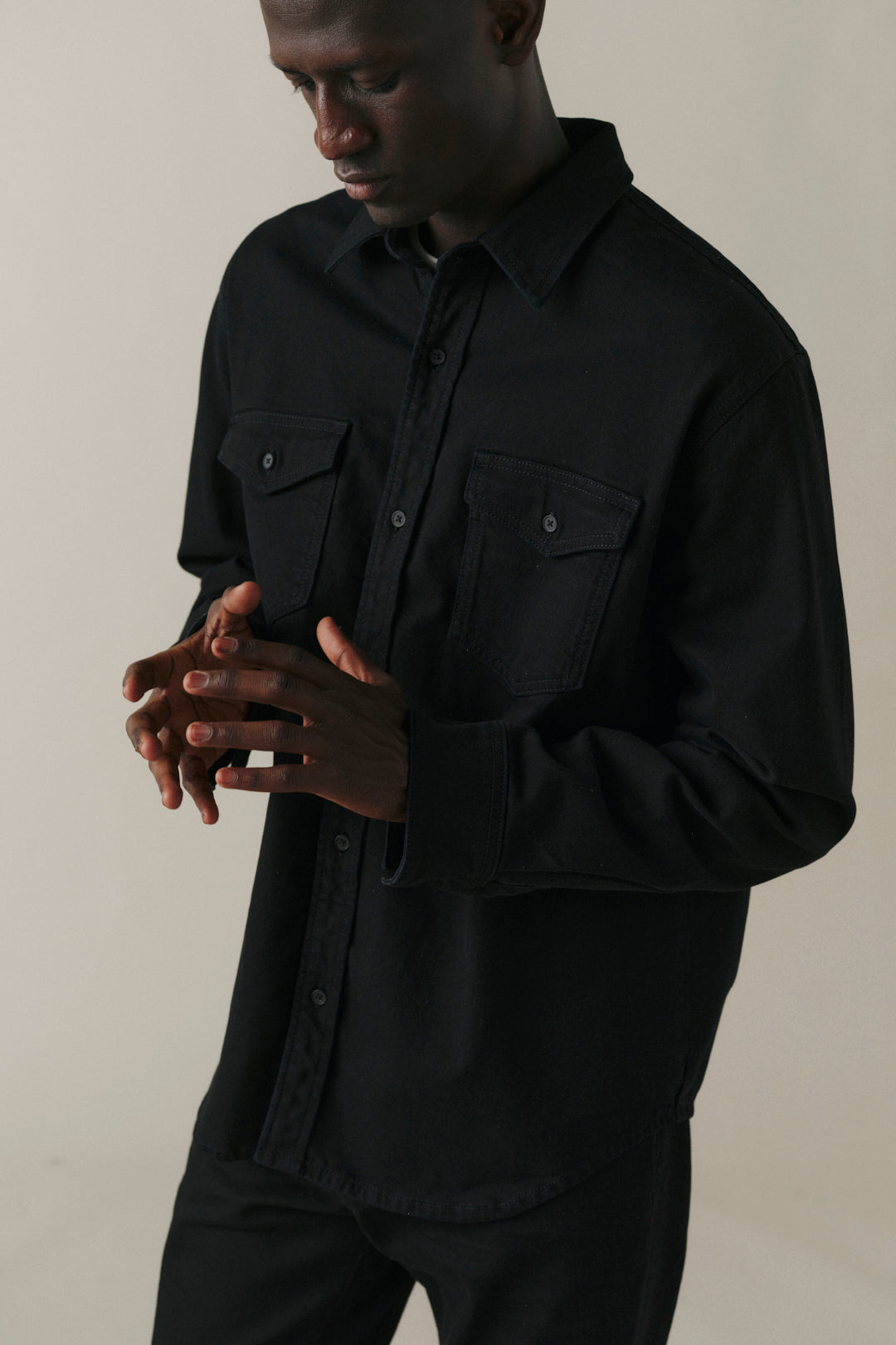 A man wearing a black long-sleeve button-up shirt with two chest flap pockets, photographed against a plain light beige studio background.