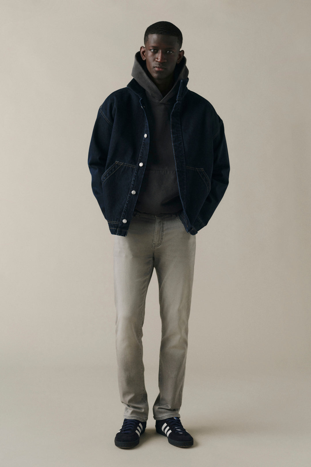 A male model stands against a plain beige studio background wearing a dark navy hooded jacket over a dark hoodie, light gray pants, and dark navy sneakers with white stripes.