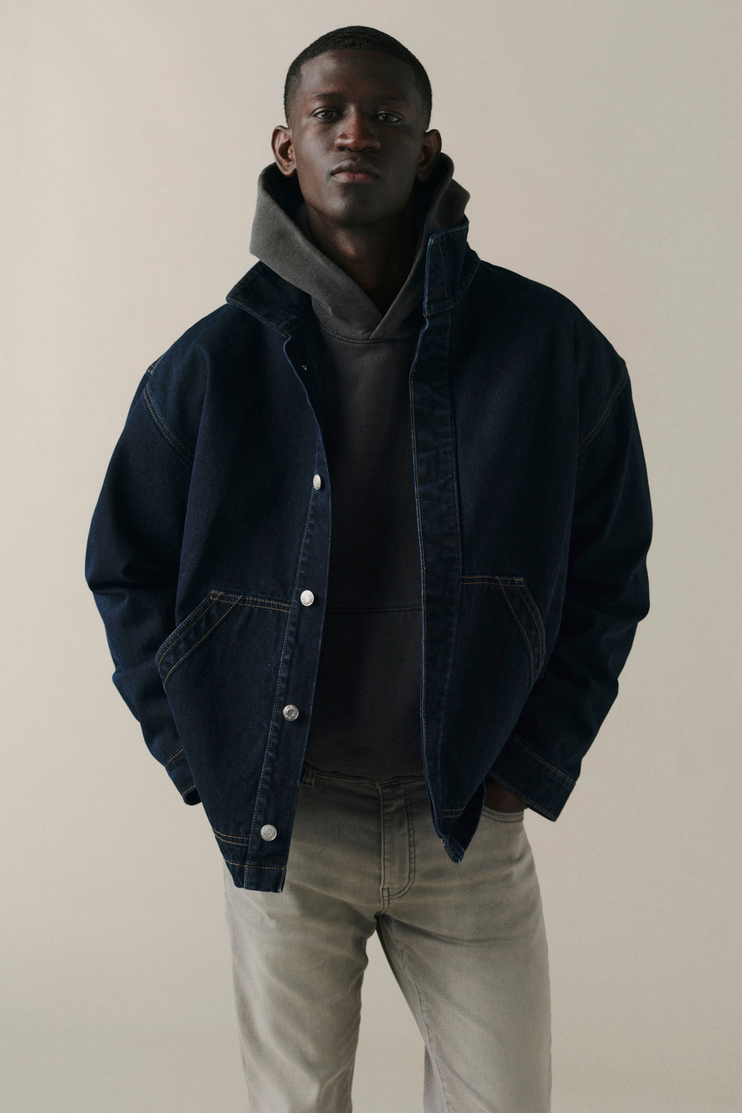 A man wearing a dark navy blue denim jacket over a dark gray hoodie and light gray jeans, standing against a plain beige studio background.