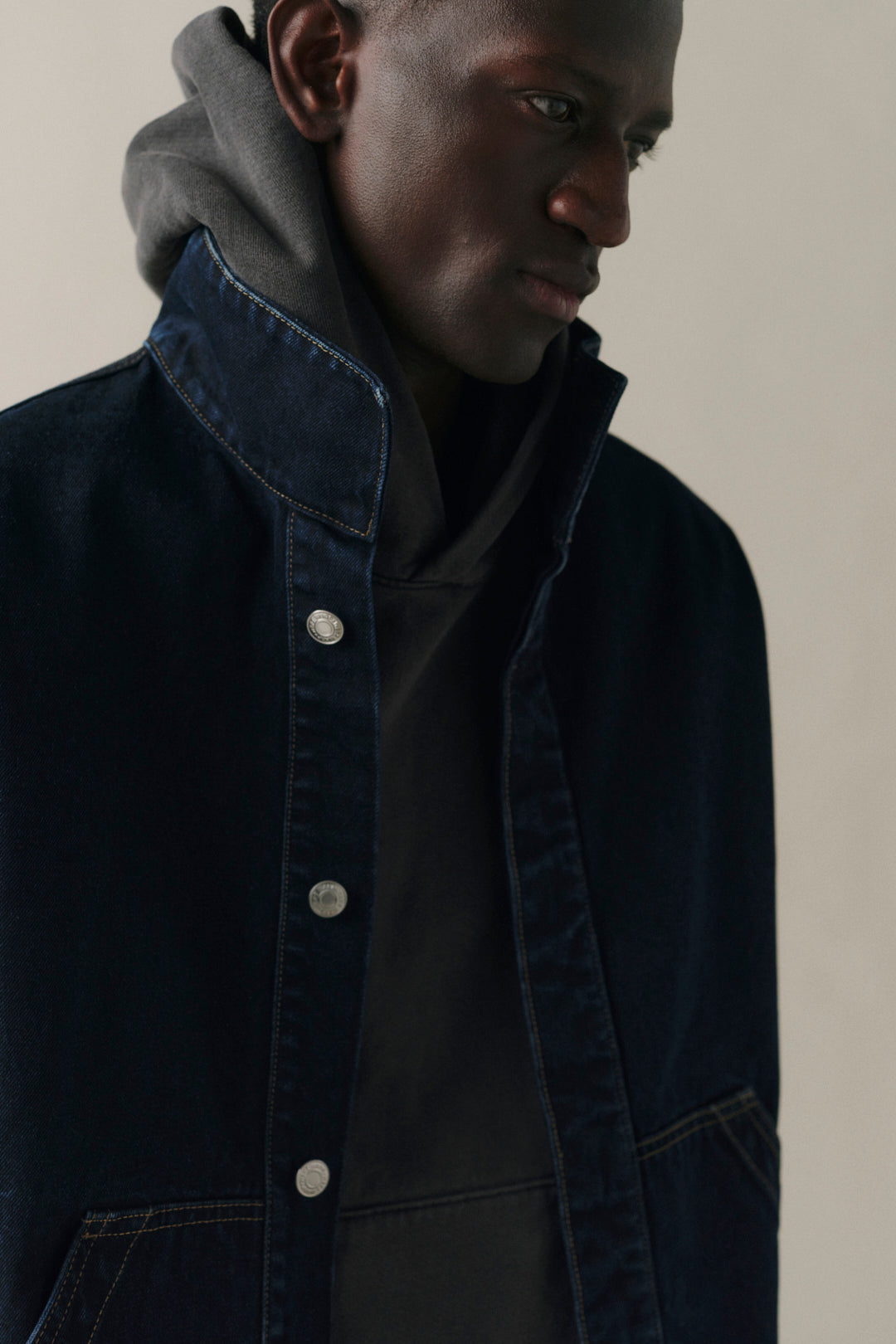 A close-up of a model wearing a dark navy blue denim jacket over a black shirt and a gray hoodie, set against a plain beige background.