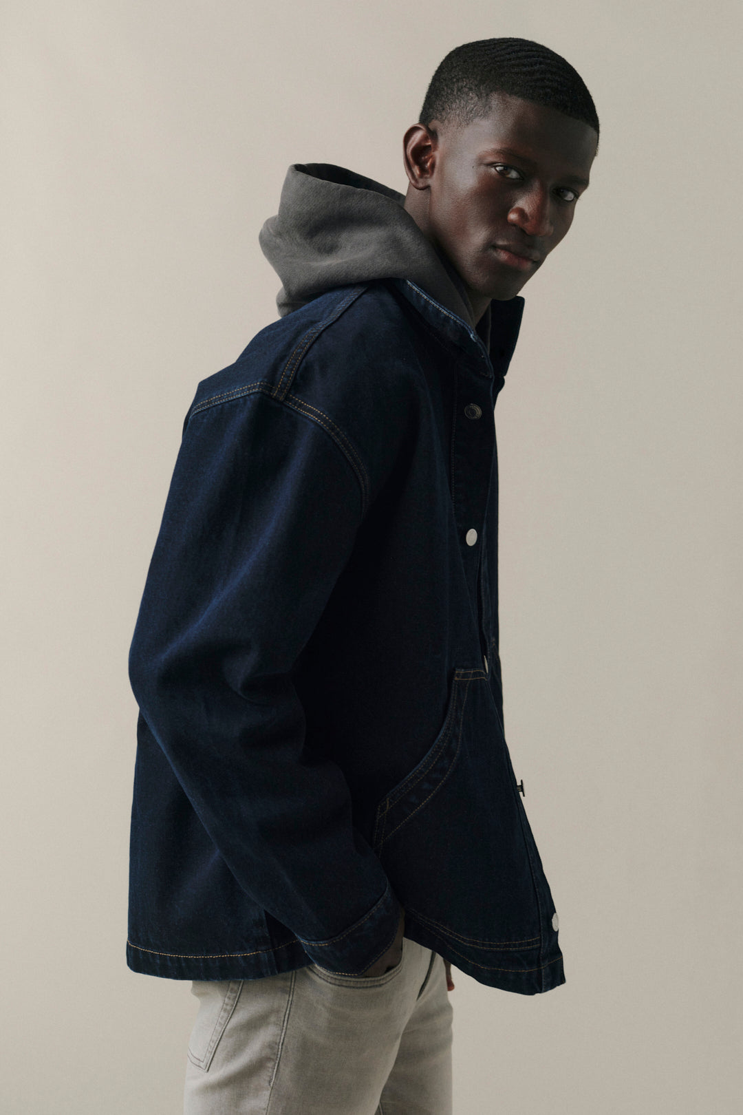 A man wearing a dark navy blue denim jacket with a gray hood, layered over a black hoodie, paired with light beige pants, set against a plain beige studio background.