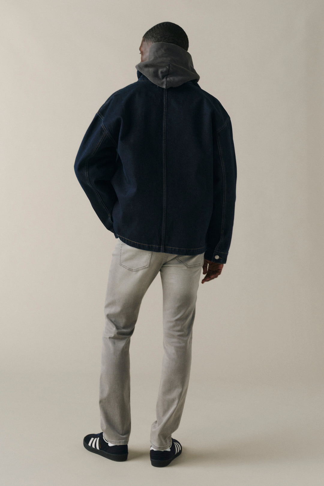 A man shown from the back wearing a dark navy zip-up jacket with a gray hood, light gray pants, and black slip-on shoes, standing against a plain beige studio background.
