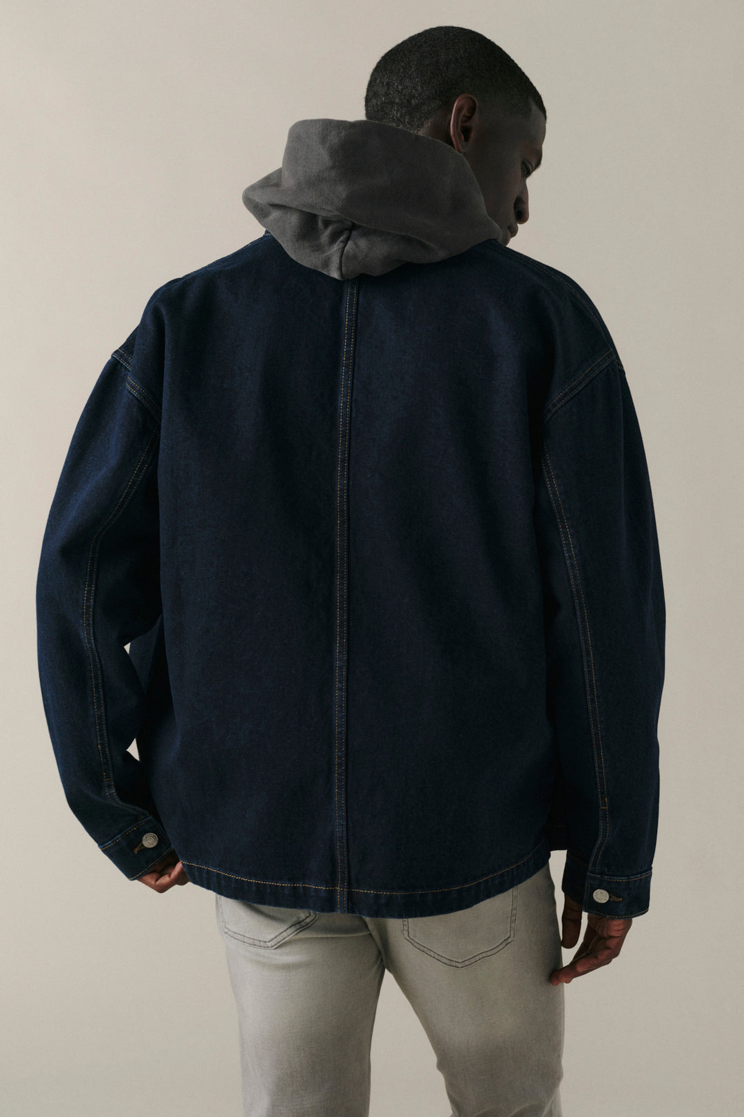 A man viewed from behind wearing a dark navy blue hooded jacket with a gray hood, paired with light gray jeans, against a plain light beige background.
