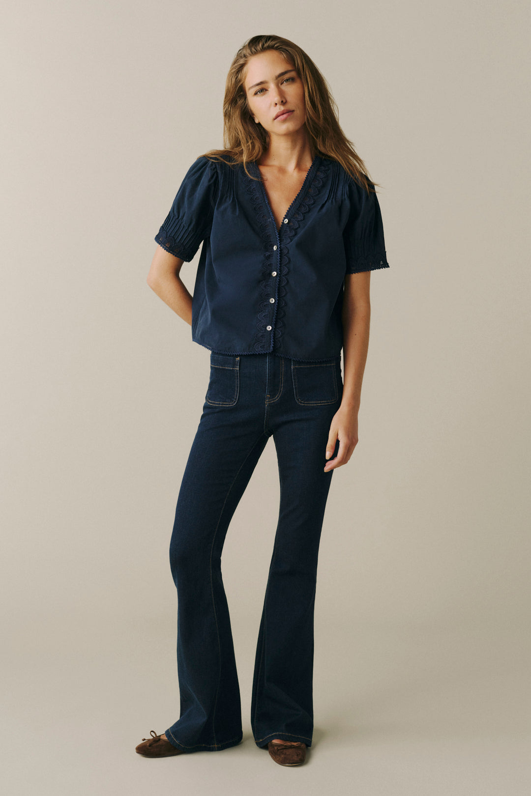 A woman modeling a dark navy blue short-sleeve button-front blouse with ruffled trim and dark blue flared jeans, standing against a plain light beige studio background.