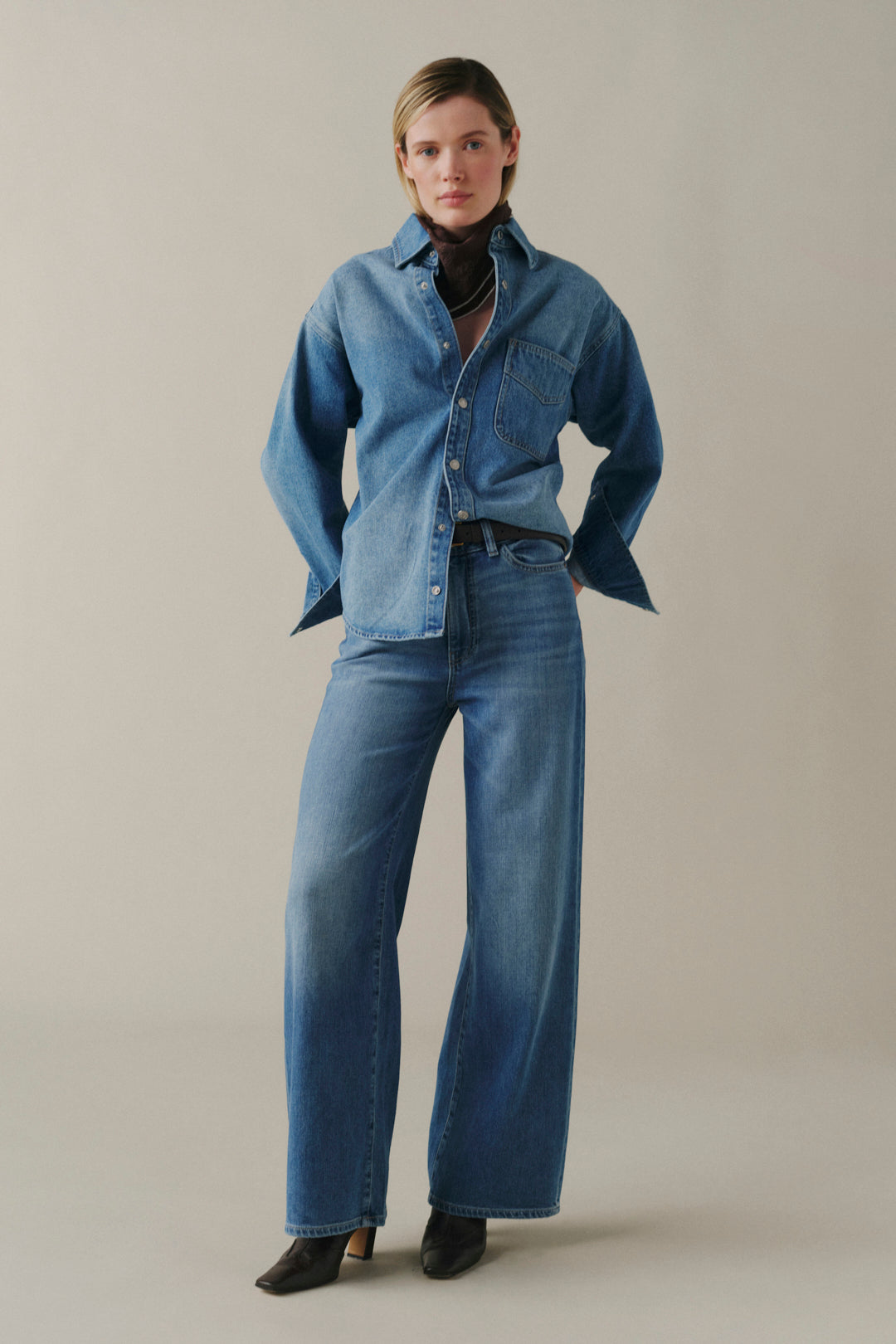 A model stands against a plain beige studio background wearing a blue denim button-up shirt, matching wide-leg jeans, a black turtleneck, and black pointed shoes.