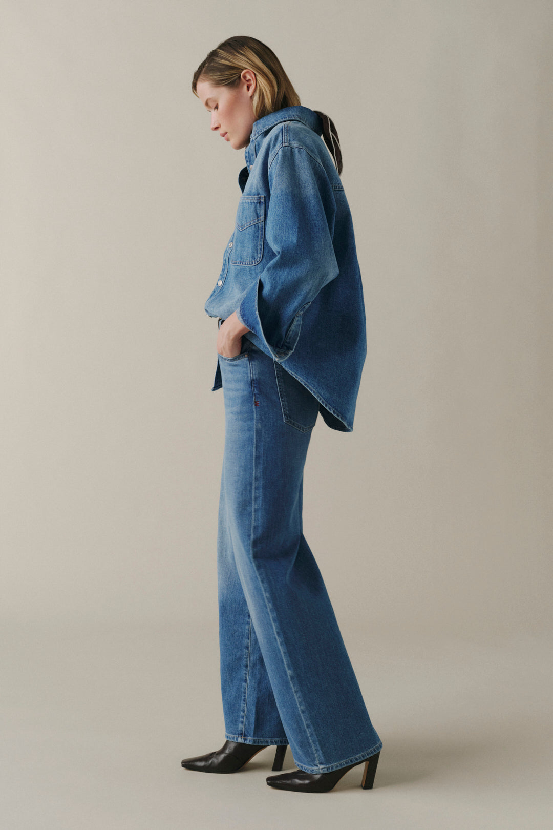 A model wearing a blue denim jacket and matching wide-leg flare jeans, paired with black pointed-toe heels, standing in profile against a neutral beige studio background.
