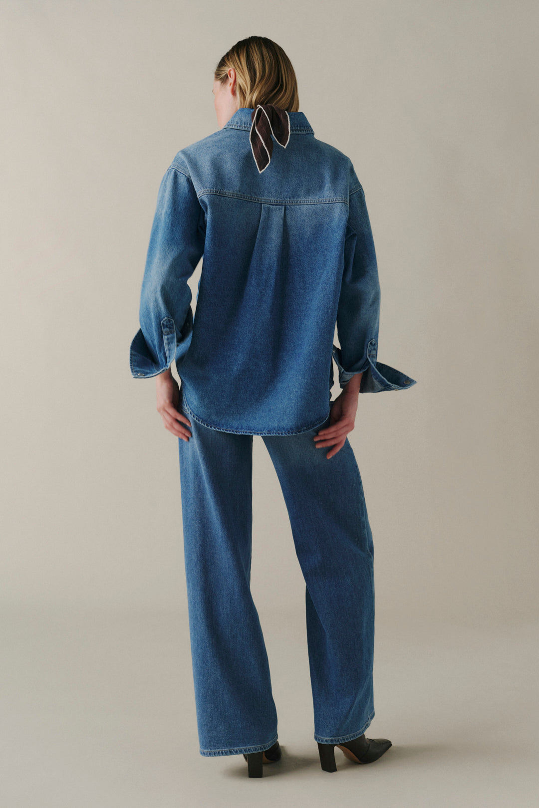 A model viewed from the back wearing a blue denim shirt and matching wide-leg jeans, standing against a neutral beige studio background.