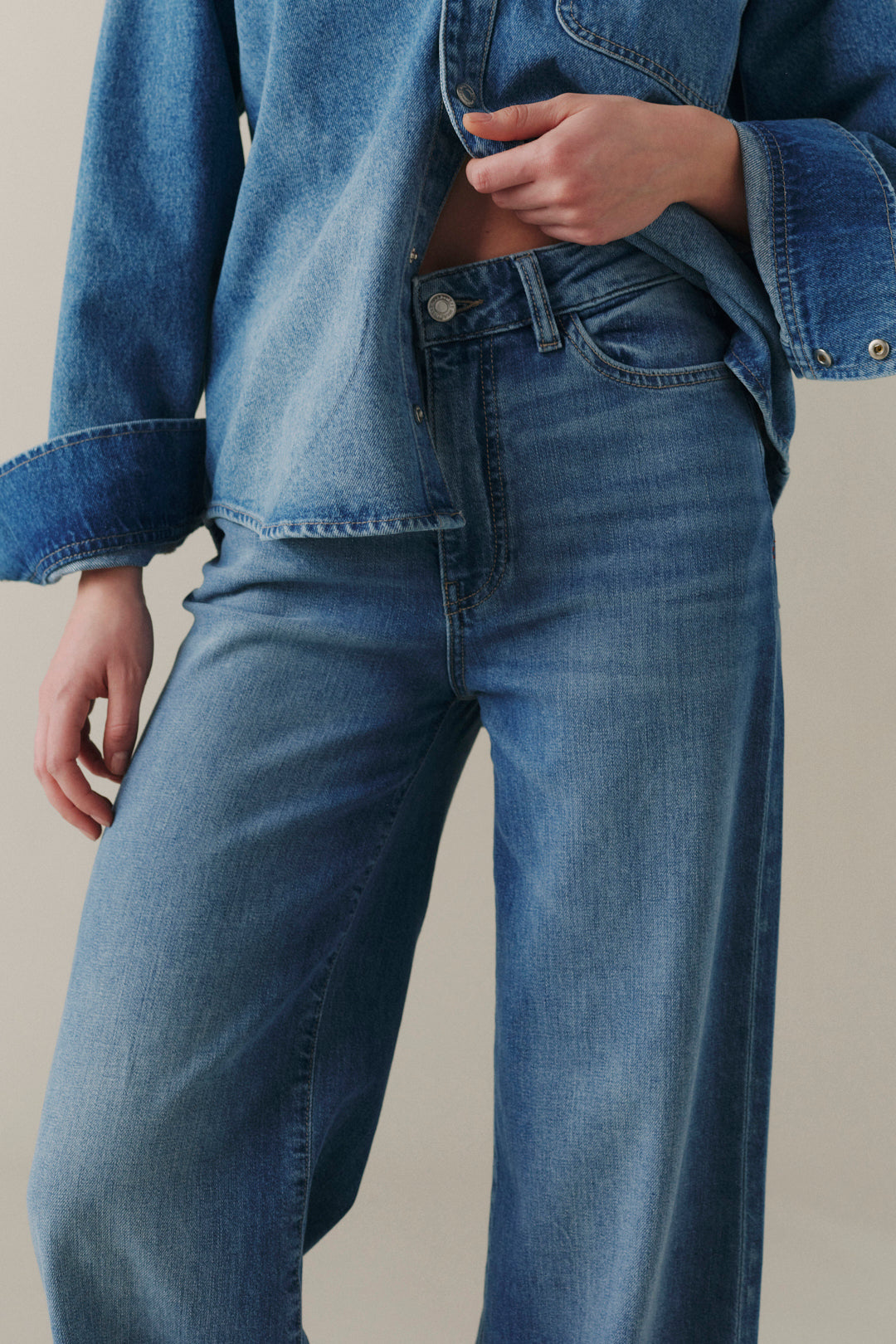 A close-up of a person wearing blue denim jeans and a matching blue denim jacket, cropped from the chest down against a plain light beige studio background.