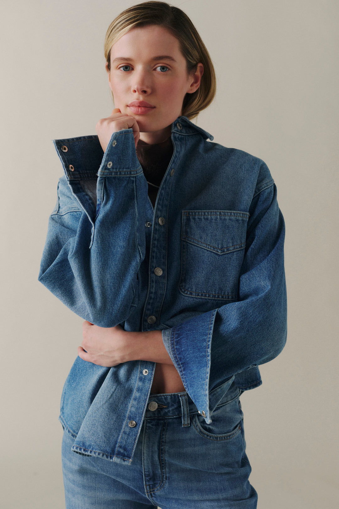 A woman wearing a cropped blue denim shirt jacket with wide sleeves and matching blue jeans, posed against a plain beige studio background.