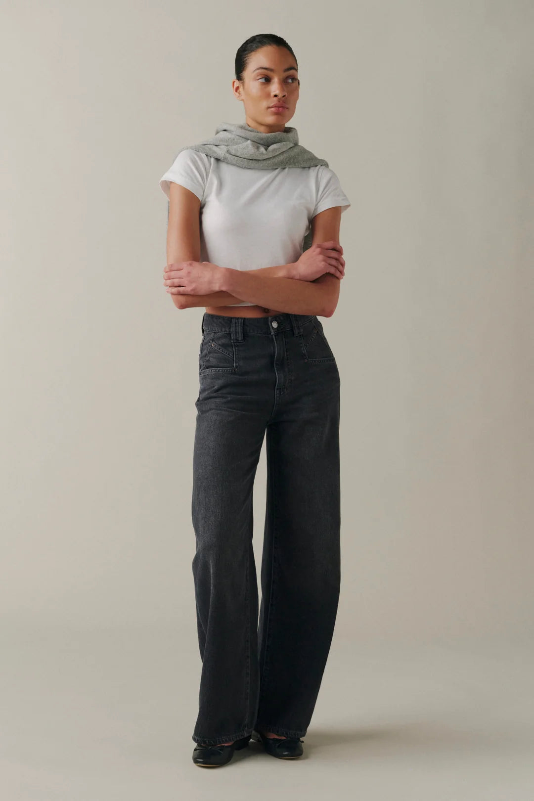 A model stands with arms crossed wearing dark charcoal wide-leg jeans, a white short-sleeve crop top, and a light gray scarf, against a plain beige studio background.