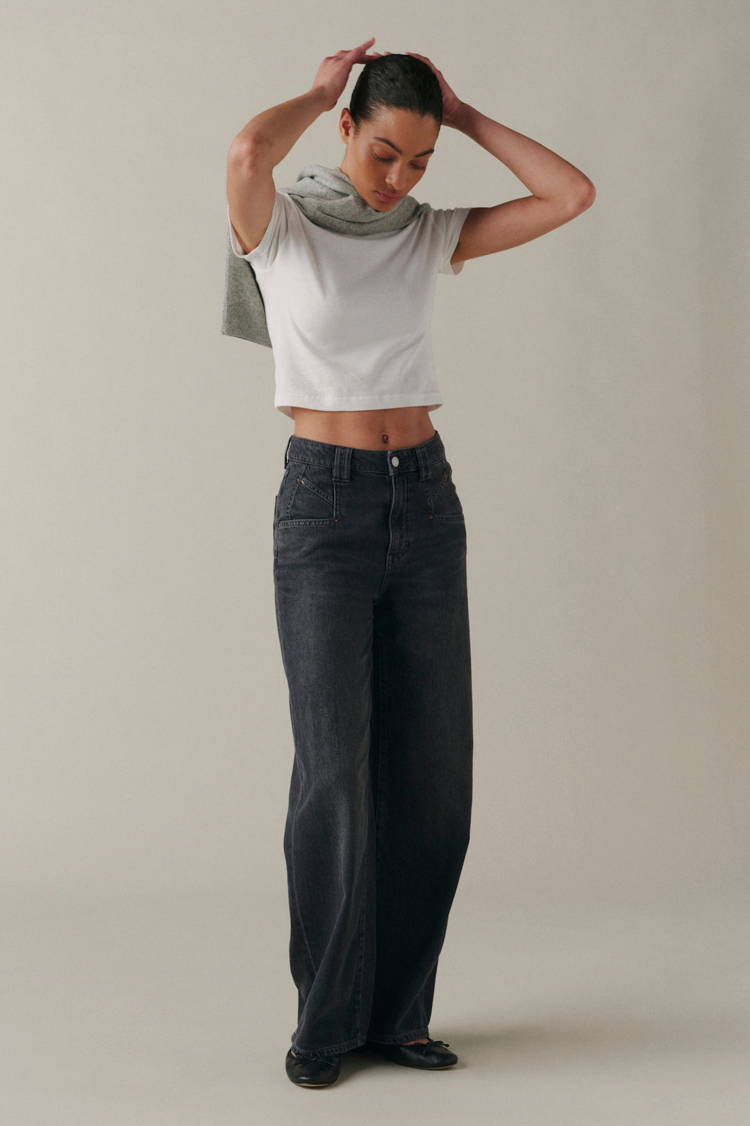 A model wearing dark gray wide-leg jeans, a white cropped t-shirt, and a light gray knit scarf, standing against a neutral beige studio background.