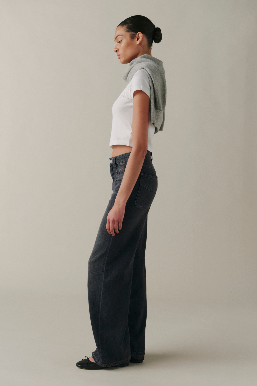 A model in profile wearing dark wide-leg jeans, a white cropped t-shirt, and a gray knit sweater draped over her shoulders, set against a plain light gray studio background.