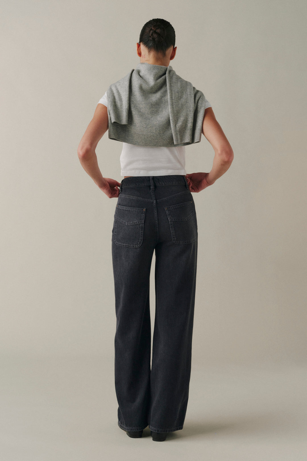 A model shown from the back wearing dark wide-leg denim jeans, a white t-shirt, and a gray cropped knit top draped over their shoulders, set against a plain light gray studio background.