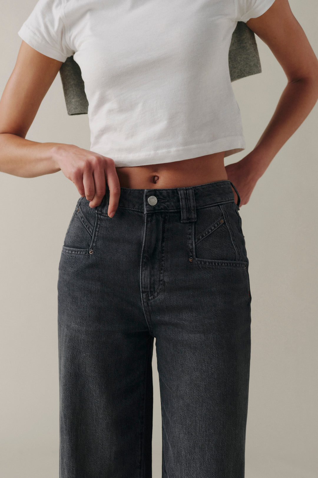 A person wearing dark gray wide-leg jeans and a white cropped T-shirt, shown from the chest down against a plain light beige studio background.
