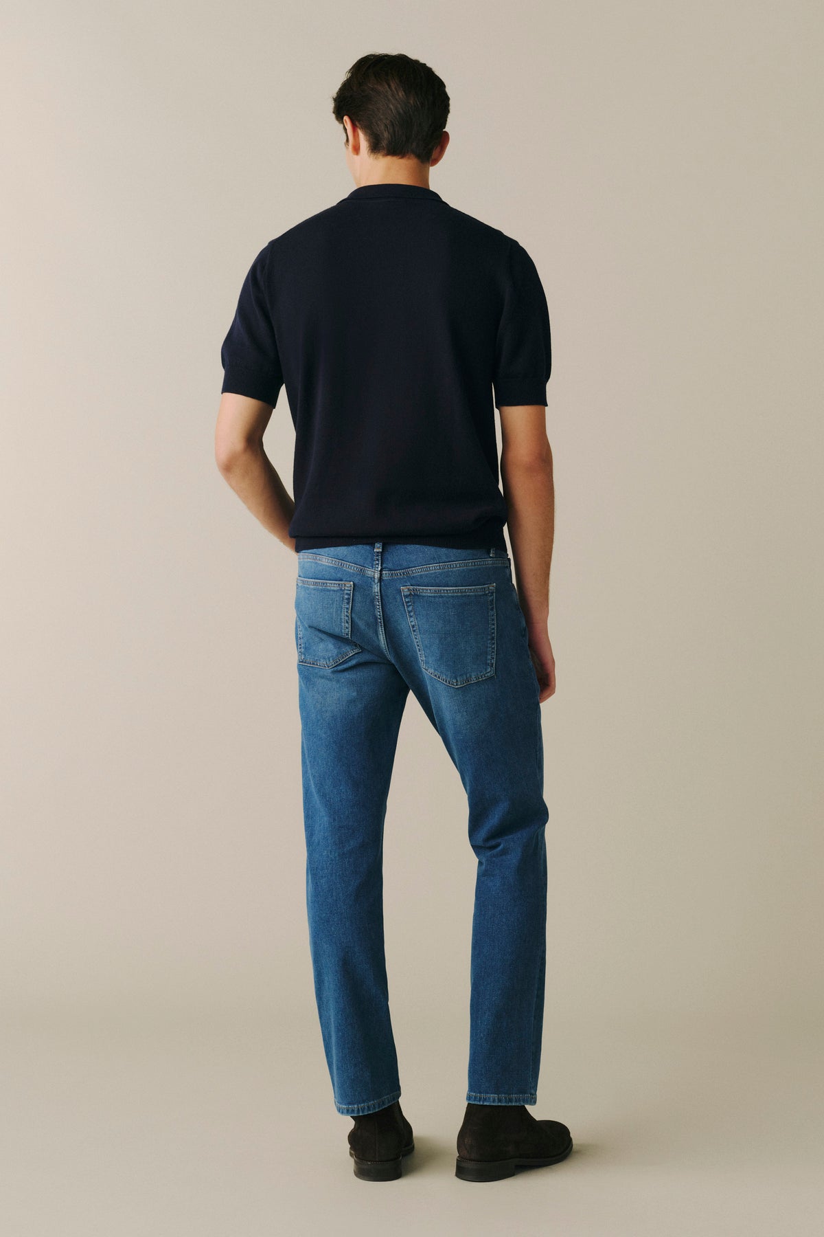 A man viewed from behind wearing a dark navy short-sleeve t-shirt, blue denim jeans, and black shoes, standing against a solid light beige background.