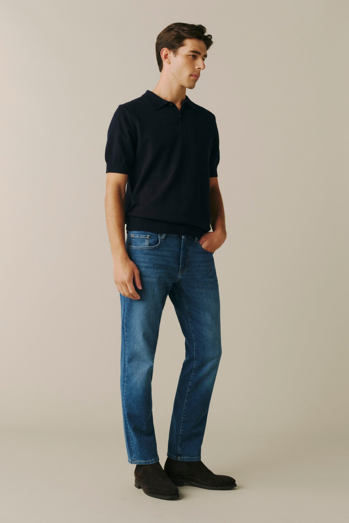 A man wearing a dark navy short-sleeve polo shirt, blue jeans, and dark brown shoes, standing against a solid light beige background.