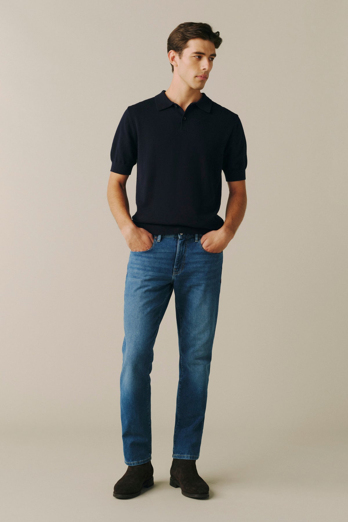 A man modeling a dark navy short-sleeve polo shirt paired with blue jeans and dark brown shoes, standing against a plain beige studio background.