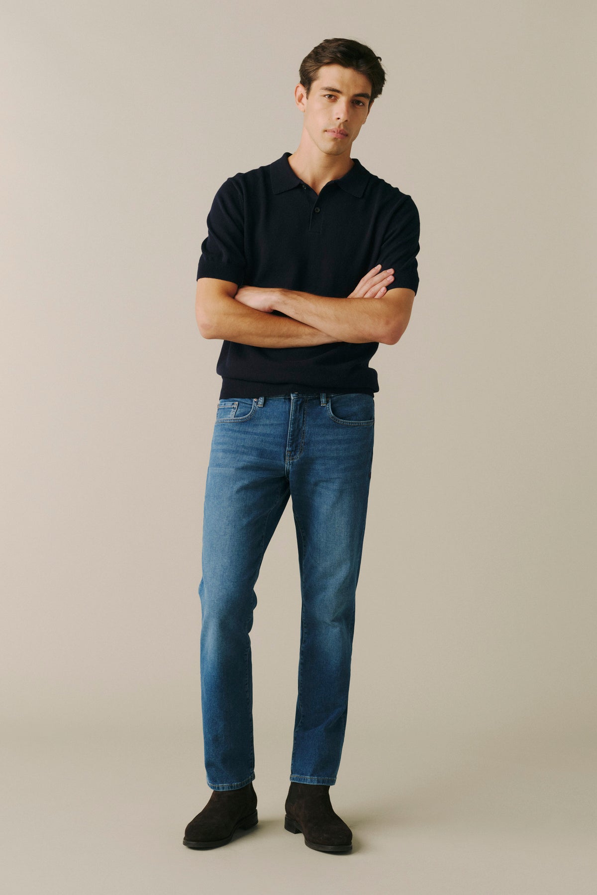 A man wearing a black short-sleeve polo shirt, blue jeans, and dark brown suede boots, standing with arms crossed against a solid beige studio background.