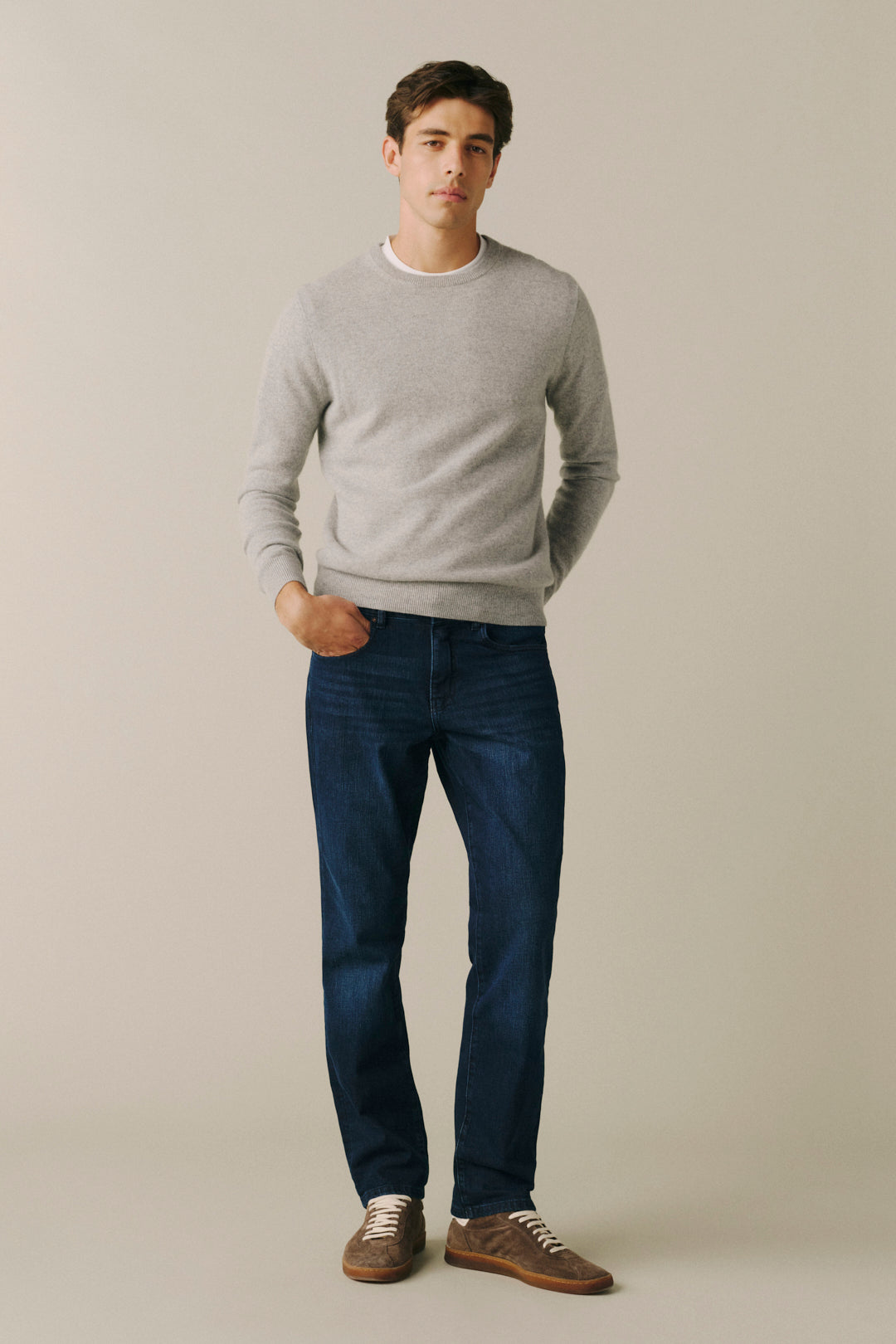 A man wearing a light gray crewneck sweater, dark blue jeans, and brown suede shoes, standing against a solid light beige studio background.