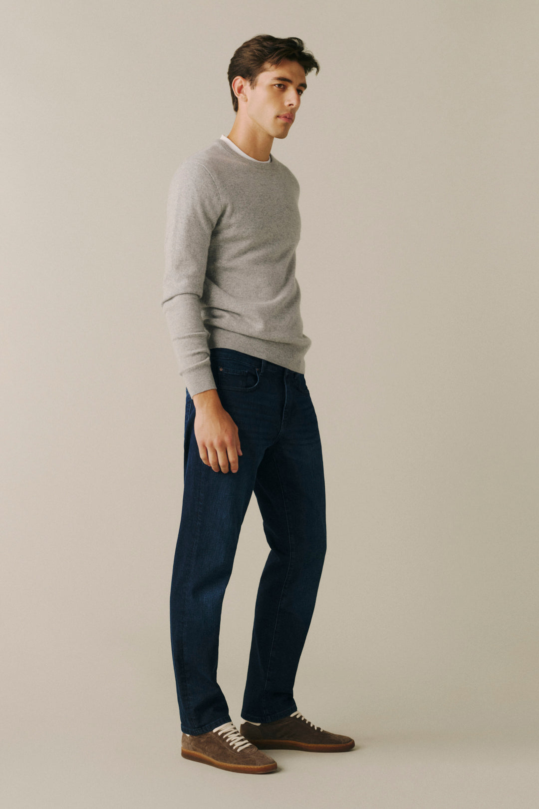 A man wearing a light gray long-sleeve crewneck sweater, dark blue jeans, and brown suede shoes, standing in profile against a solid light beige background.