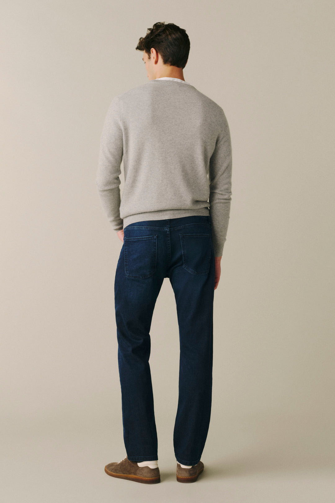 A man viewed from behind wearing a light gray knit sweater, dark blue jeans, and brown suede shoes, standing against a plain beige studio background.
