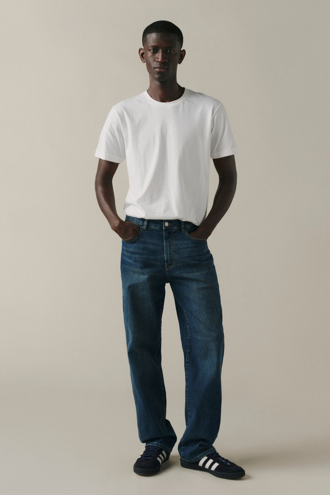 A man modeling a white crew-neck t-shirt, dark blue jeans, and black sneakers with white stripes, standing against a solid beige studio background.