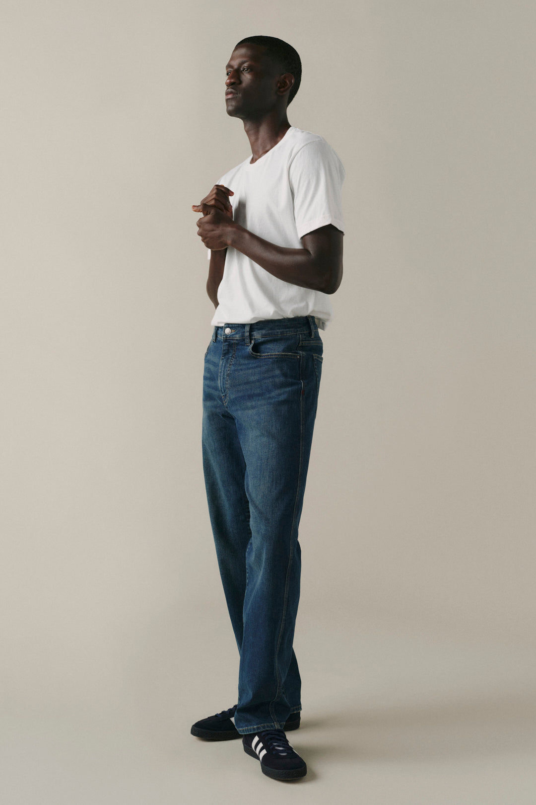 A man wearing a white t-shirt, blue jeans, and black sneakers stands in profile against a plain beige studio background.