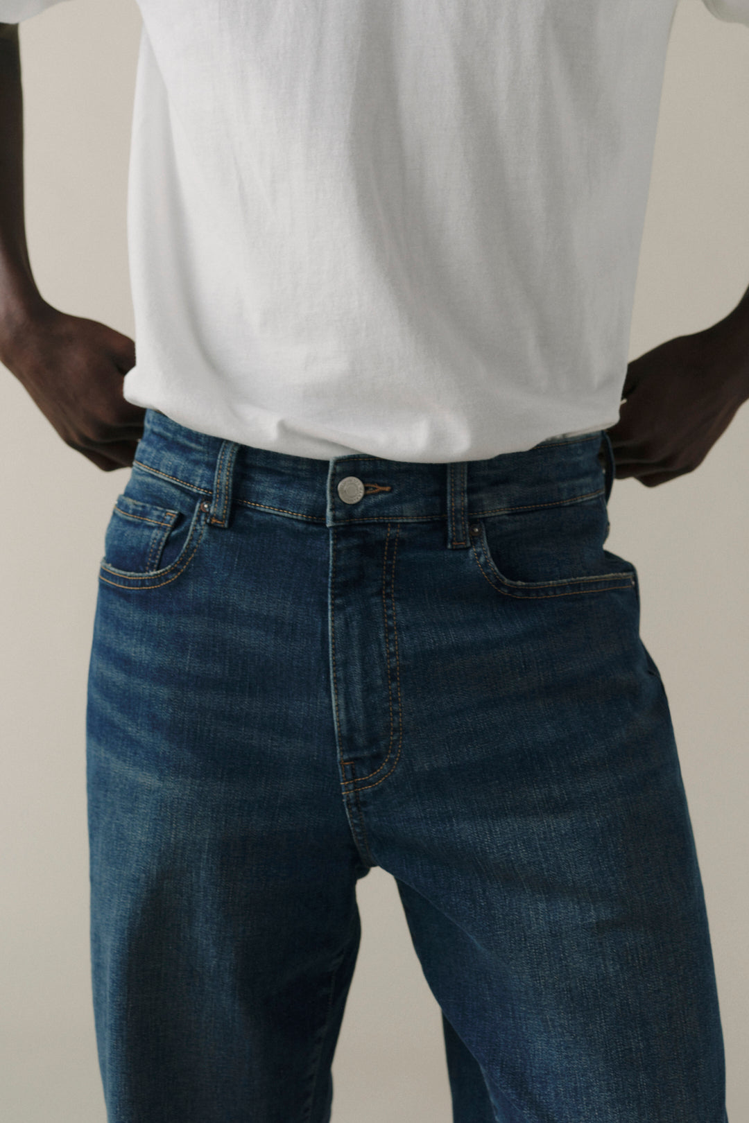 A close-up of a person wearing dark blue denim jeans and a white t-shirt, with hands in pockets, against a plain light beige background.