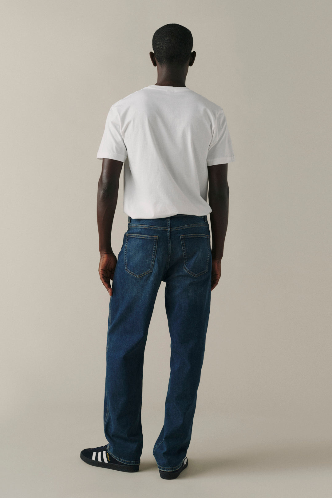A man viewed from behind wearing a white t-shirt, dark blue jeans, and black slide sandals with white stripes, standing against a solid light beige background.